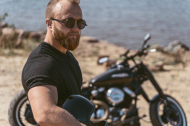 Close up of a handsome man holding helmet and standing next to his bike - Фото, зображення