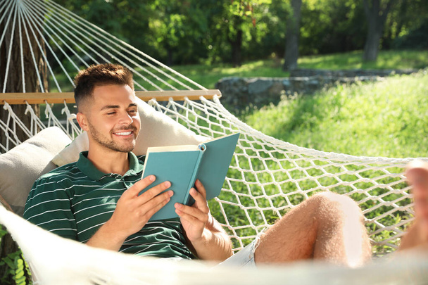 Young man reading book in comfortable hammock at green garden - Foto, afbeelding