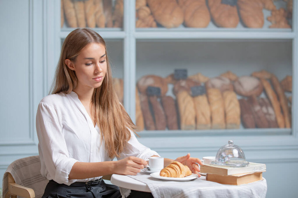 Woman is heaving breakfast with croissant and coffee in the morning. Very beautiful girl with long blonde hair having lunch in a bakery - Foto, imagen