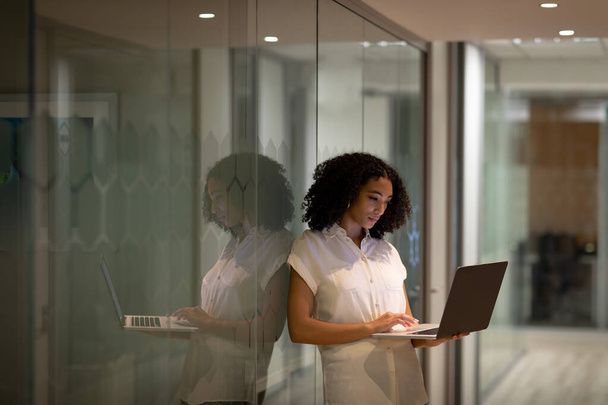 Widok z przodu młoda mieszana rasa profesjonalistka pracująca do późna w nowoczesnym biurze, stojąca w korytarzu za pomocą laptopa, odbita w szklanej ścianie - Zdjęcie, obraz
