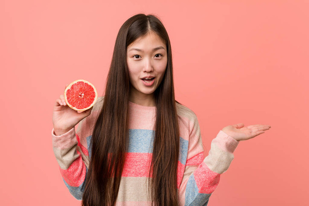 Молодая азиатка с грейпфрутом произвела впечатление, держа пространство для копирования на ладони
. - Фото, изображение