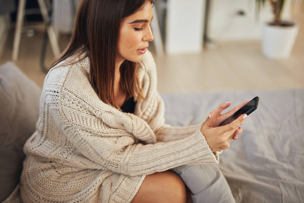 Atrakcyjna biała brunetka ubrana w beżowy sweter, siedząca na łóżku w sypialni i korzystająca ze smartfona. - Zdjęcie, obraz
