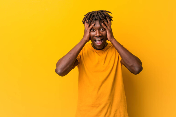 Jonge zwarte man met rasta 's over gele achtergrond lacht vrolijk handen op het hoofd. Geluksconcept. - Foto, afbeelding