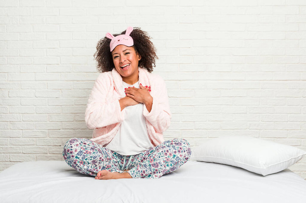 Mujer afroamericana joven en la cama con pijama tiene expresión amistosa, presionando la palma en el pecho. Concepto de amor
. - Foto, imagen
