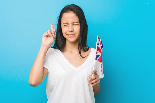 junge hispanische Frau, die die Fahne des Vereinigten Königreichs hält und die Daumen drückt, um Glück zu haben - Foto, Bild