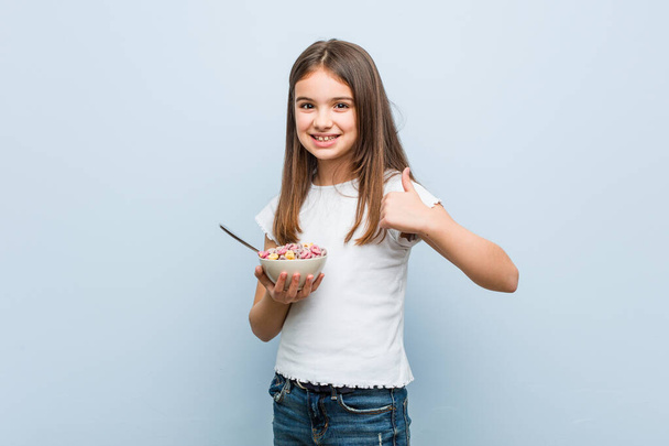 Pequeña chica caucásica sosteniendo un tazón de cereal sonriendo y levantando el pulgar
 - Foto, Imagen