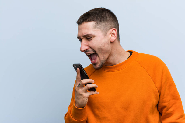Joven hombre caucásico sosteniendo un teléfono gritando emocionado al frente
. - Foto, Imagen
