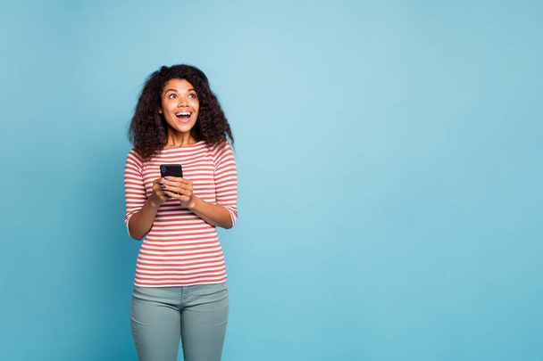 Photo of cheerful charming cute nice curly wavy black woman holding telephone rejoicing with feedback being got wearing striped t-shirt trousers pants looking into empty space isolated blue pastel - Foto, Bild