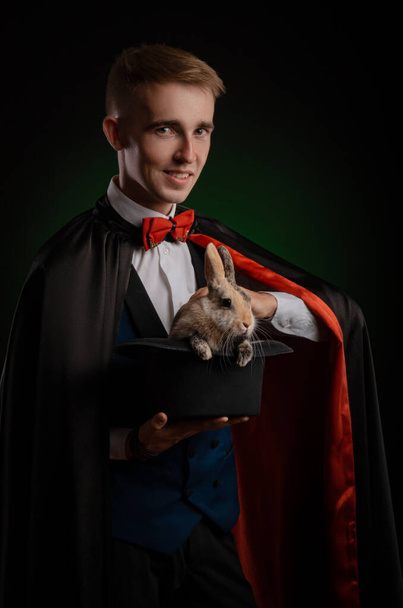 un joven mago ilusionista sosteniendo un sombrero con un conejo - Foto, Imagen