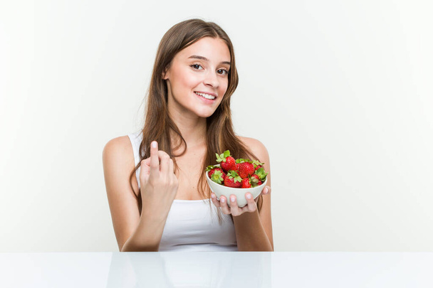 Young caucasian woman holding a strawberries bowl pointing with finger at you as if inviting come closer. - Photo, Image
