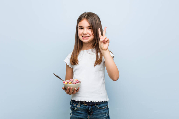 Pequeña chica caucásica sosteniendo un tazón de cereal mostrando el número dos con los dedos
. - Foto, Imagen