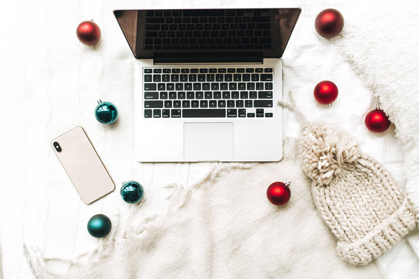 Christmas / New Year composition. Flat lay, top view. Laptop and cellphone lying on the white bed with white blanket decorated with christmas red and blue balls and winter knitted hat. Minimal business freelance concept. - Foto, Bild