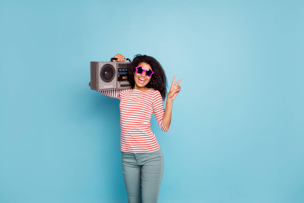 Photo of funny dark skin lady show v-sign hold cool vintage recorder on shoulder ready start party wear sun specs casual clothes isolated pastel blue color background - Foto, imagen