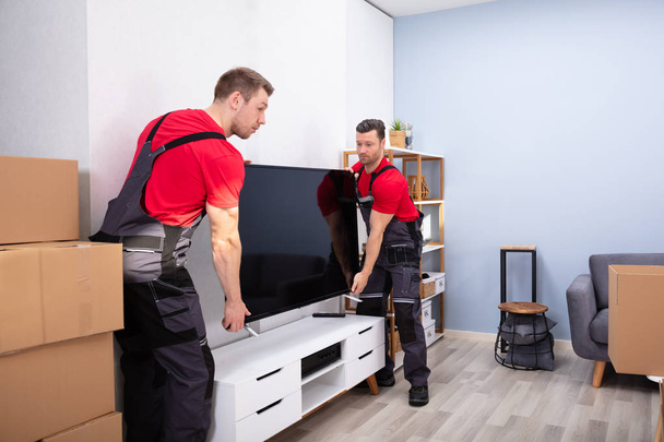 Two Male Professional Movers In Uniform Unloading LCD Television In The Living Room - Φωτογραφία, εικόνα