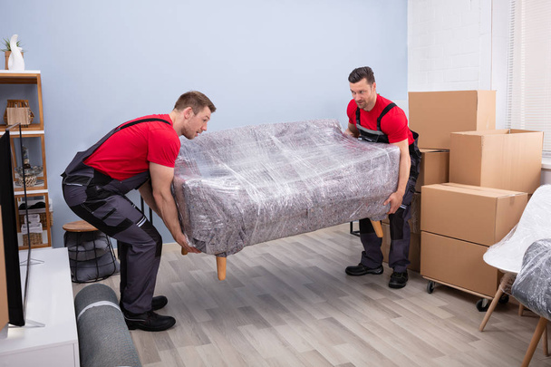 Portrait Of Two Young Happy Male Movers Carrying Wrapped Sofa In New House - Foto, Imagen