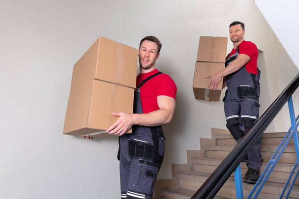 Portrait Of A Young Male Movers In Uniform Carrying Cardboard Boxes Walking Downward On Staircase - Fotografie, Obrázek