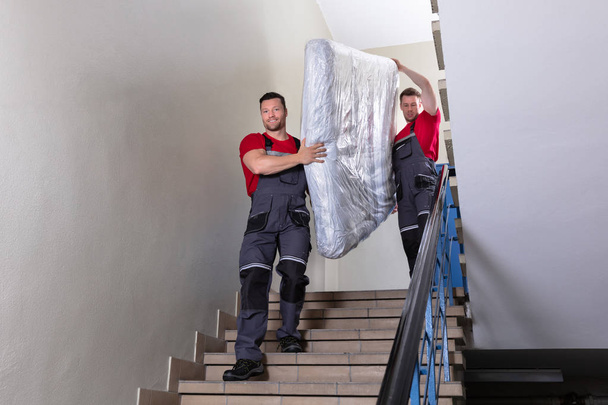 Two Young Male Movers In Uniform Carrying The Wrapped Mattress While Moving Downward The Staircase - Foto, afbeelding