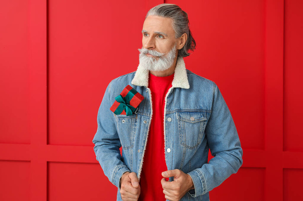 Portrait of stylish Santa Claus with Christmas box on color background - Fotó, kép