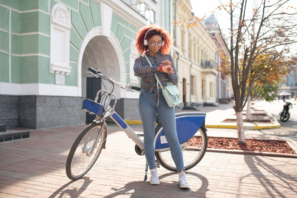 Girl near bike. Curly red-haired girl looking at notifications on smart watch standing near bike - 写真・画像