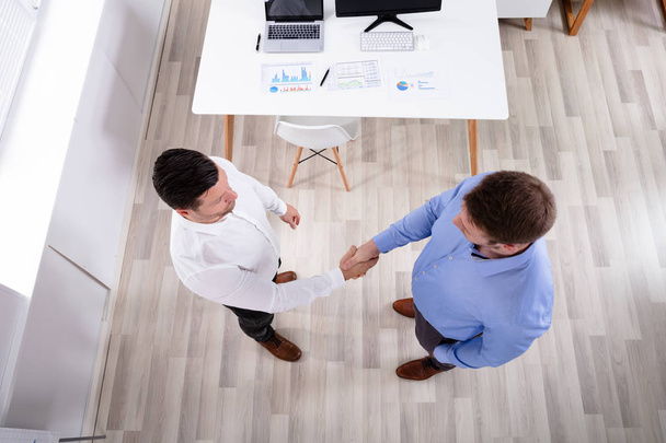 Возвышенный вид двух бизнесменов, стоящих в офисе и пожимающих друг другу руки
 - Фото, изображение
