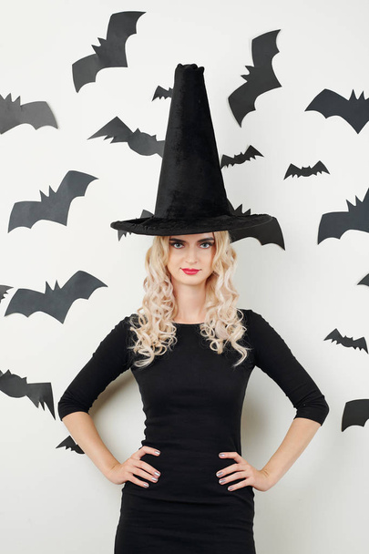 Retrato de joven bruja confiada de pie contra la pared blanca con pegatinas de murciélagos negros
 - Foto, Imagen