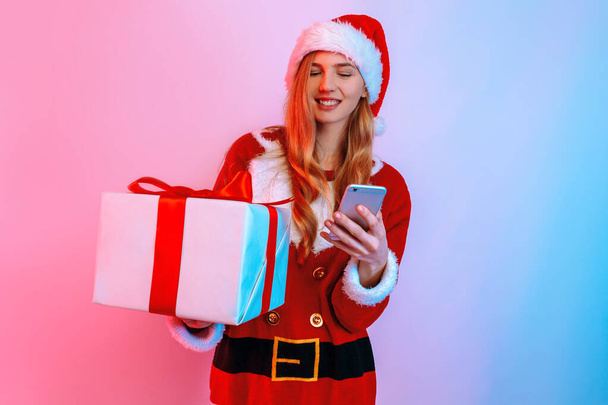 Happy young woman in Santa hat, with Christmas gifts, uses smartphone - Foto, Bild