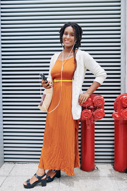 Портрет африканської стилістки в елегантному оранжевому вбранні посміхається перед камерою, слухаючи музику на її мобільному телефоні. - Фото, зображення