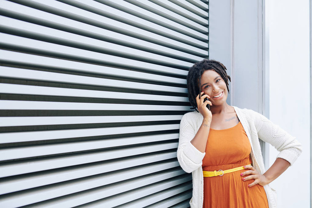 Retrato de mujer africana alegre en ropa de moda de pie hablando por teléfono móvil y sonriendo a la cámara contra la pared rayada en la ciudad
 - Foto, imagen