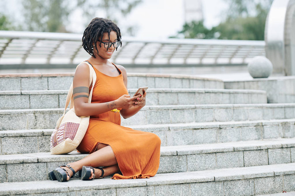 Αφρικανική όμορφη γυναίκα κάθεται στις σκάλες πληκτρολογώντας ένα μήνυμα στο κινητό της τηλέφωνο και χαμογελώντας σε εξωτερικούς χώρους - Φωτογραφία, εικόνα