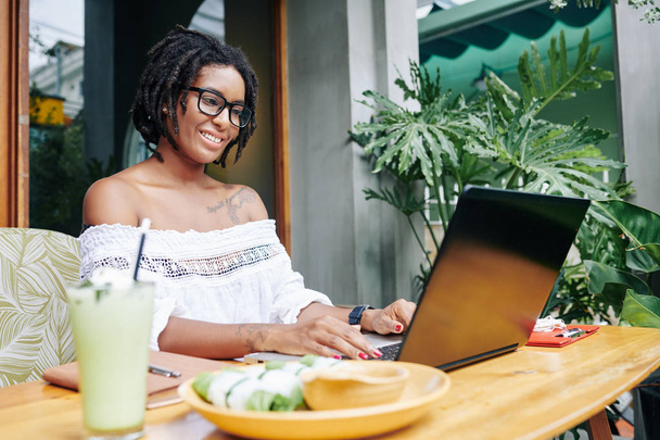 African smiling businesswoman in eyeglasses sitting at the table with food and typing on laptop computer in outdoor cafe - Фото, зображення