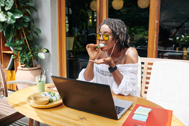 African young woman in sunglasses sitting at wooden table in front of laptop and eating during her work in outdoor cafe - Photo, Image