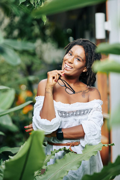 Portret afrykańskiej pięknej kobiety trzymającej okulary i uśmiechającej się do aparatu fotograficznego w naturze - Zdjęcie, obraz