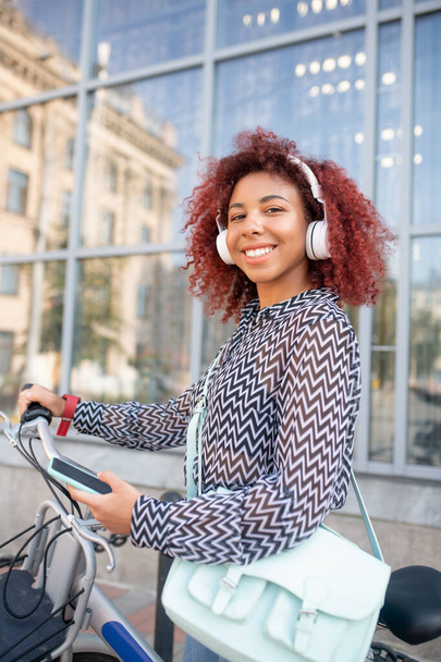 Curly student. Curly beaming red-haired student wearing earphones standing near bike - Photo, Image