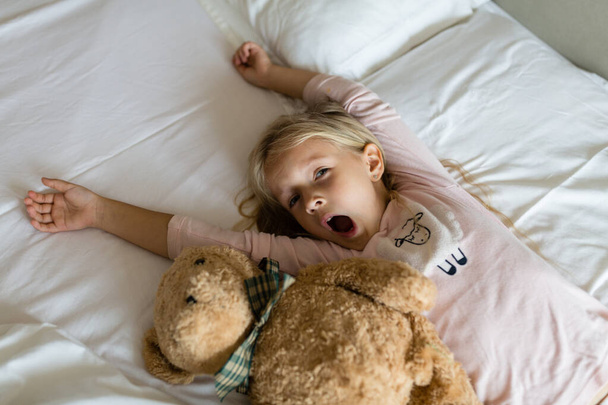 Κάτοψη του χαριτωμένου ξανθού κοριτσιού με πιτζάμες και δεμένα μάτια ξαπλωμένο σε λευκό κρεβάτι με αρκουδάκι, ξυπνώντας νωρίς το πρωί πριν πάτε στο νηπιαγωγείο. Ώρα για ύπνο, αφύπνιση και χαλάρωση - Φωτογραφία, εικόνα
