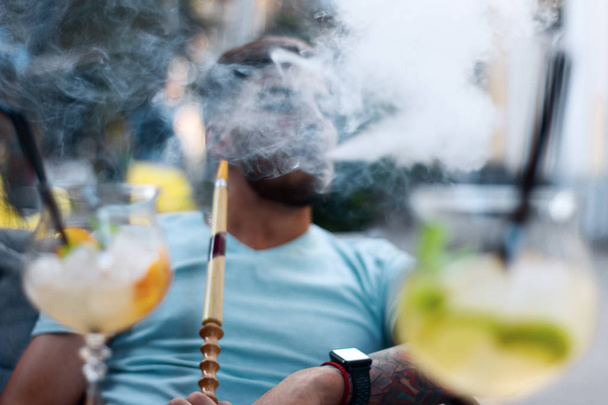 Όμορφος τύπος εκπνέει καπνό ναργιλέ. Νεαρός άντρας καπνίζει ένα αρωματικό ανατολίτικο ναργιλέ έξω. Υπόλοιπο με hookah και κοκτέιλ στην περιοχή του καλοκαιριού. - Φωτογραφία, εικόνα