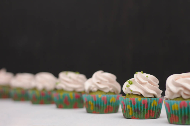 Groene cupcakes met roomhoed in veelkleurig papier op een witte tafel op een donkere achtergrond. Focus op cake, scherptediepte effect - Foto, afbeelding