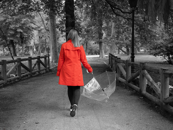 Μια λευκή κοπέλα με κόκκινη καμπαρντίνα διασχίζει ένα πάρκο με μια διάφανη ομπρέλα στο χέρι της.. - Φωτογραφία, εικόνα