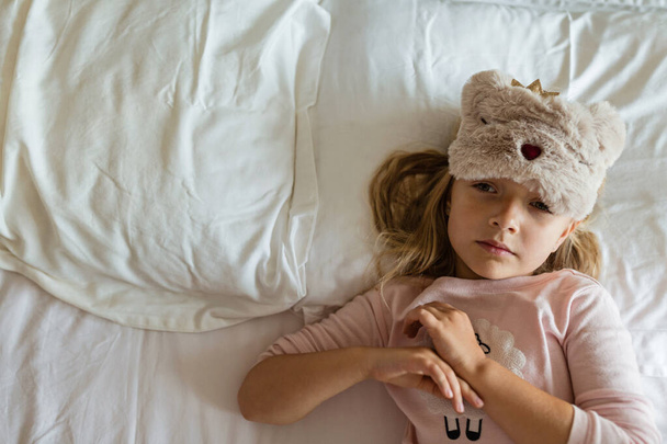 Top view of cute blonde little girl in pajamas and blindfold lying in white bed, awaking early in the morning before going to kindergarten. Bedtime, awakening and relaxation concept - Foto, Bild