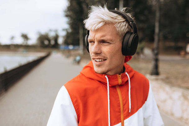 Outdoor close-up portrait of handsome young man enjoying weather in city park, waiting for his girlfriend, while listening to his favorite music from wireless headphones in the park. - Фото, изображение