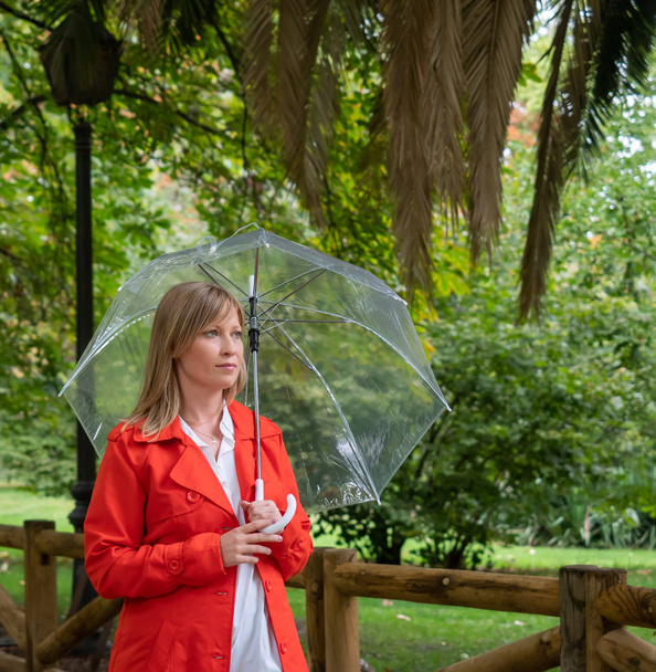 Kırmızı gabardinli beyaz kız şeffaf bir şemsiyeyle parktan geçiyor.. - Fotoğraf, Görsel