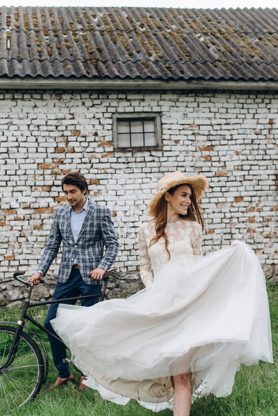 A beautiful young couple bride and groom with a bicycle near the old house - Zdjęcie, obraz