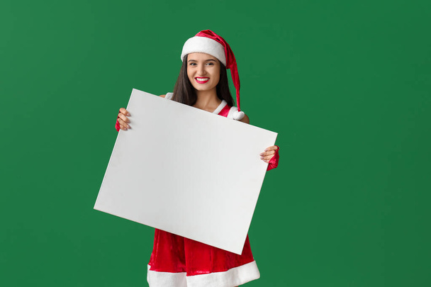 Beautiful young woman in Santa Claus costume and with blank poster on color background - Photo, Image