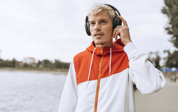 Attractive young man listening to his favorite music from wireless headphones using smart phone during walking in the park. - Photo, Image