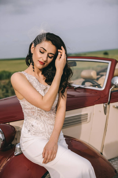 Stylish pretty bride sitting on a red retro car on a field road. - Фото, изображение