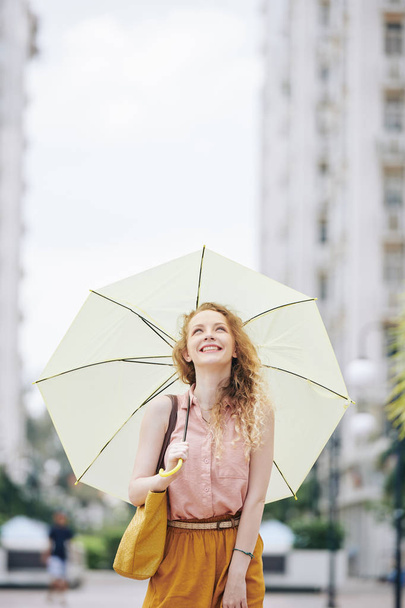 Šťastná emocionální mladá žena s blond kudrnaté vlasy kráčející po ulici s velkým deštníkem a při pohledu na obloze - Fotografie, Obrázek