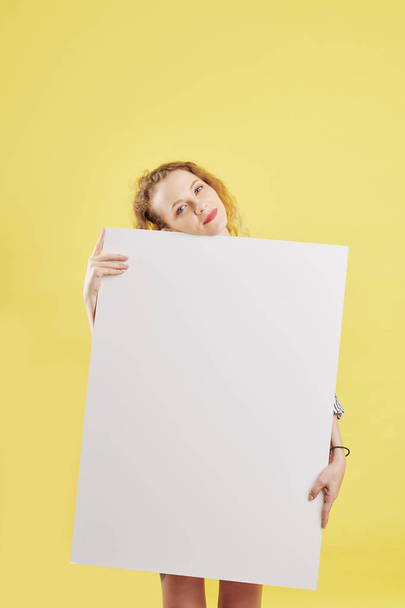Pensive smiling young woman holding big empty white paper sheet and looking at camera - Фото, изображение