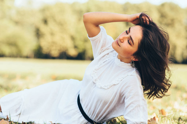 Image of attractive young woman with windy hair lying on the grass, enjoying the warm weather, wearing white dress on nature background.Pretty emale relaxing in the park on sunny day. - Photo, image