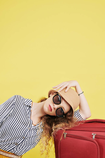 Beautiful young woman in big sunglasses resting her head on big packed suitcase and looking at camera, isolated on yellow background - Fotografie, Obrázek