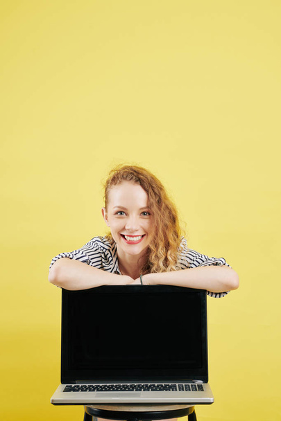 Cheerful young female entrepreneur in striped blouse leaning on opened laptop with black empty screen - Foto, Bild