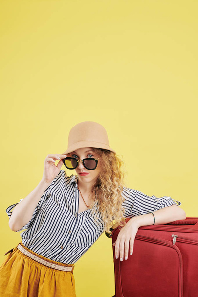 Serious young woman leaning on big packed suitcase and taking off sunglasses to look at camera - Фото, зображення
